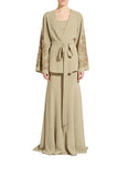 A model stands against a light gray studio background wearing a long beige wrap robe with matching wide sleeves featuring ornate gold embroidery, a tie belt at the waist, and a flowing maxi skirt.