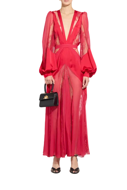 A model stands against a light gray studio background wearing a bright red sheer maxi dress with dramatic balloon sleeves, a deep V-neckline, and a high front slit. She is holding a small black handbag with a gold logo.
