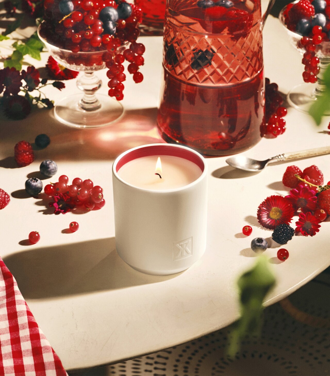 A lit candle in a white cylindrical holder with a red inner rim. The holder has an embossed logo.