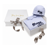 Light blue baby sleepsuit with collar and matching beanie hat. Both feature dark blue embroidered 'Prince' text with a gold-t