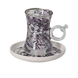Side view of a clear glass tea cup with purple floral motifs and a matching ceramic saucer.