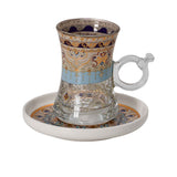 Close-up of a clear glass tea cup with handle and ornate gold/blue patterns resting on a matching saucer.