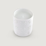 White ceramic candle holder with embossed crown, horse, and plume designs. White wax and a single wick are visible inside. Th