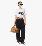 Person wearing a white t-shirt with a graphic design, black pants, and holding a brown leather bag on a white background.