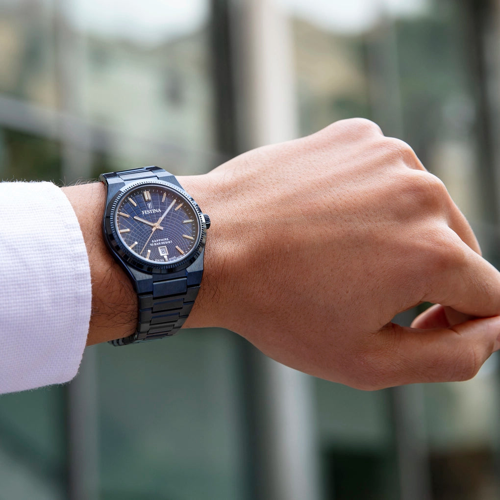 Dark blue Festina watch with a round case, textured bezel, and metal link bracelet. The blue dial has a grid pattern, gold-to