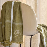 Olive green woven throw blanket with a white vertical stripe, horizontal herringbone stripes, fringed edges, and an embroider