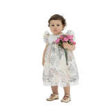 A toddler girl with curly brown hair wearing a floral print cream dress with flutter sleeves and gold Mary Jane shoes, holding a bouquet of pink peonies against a white background.
