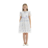 A young girl standing against a white background wearing a knee-length white lace party dress with light purple floral embroidery, sheer short sleeves, and a lavender ribbon bow at the waist.