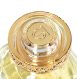 A detailed, top-down macro shot of the luxurious circular gold cap of the Touch of Oud perfume bottle. The polished gold center features an engraved brand monogram logo, surrounded by an ornate, filigree-patterned gold rim enclosed in clear, heavy acrylic.